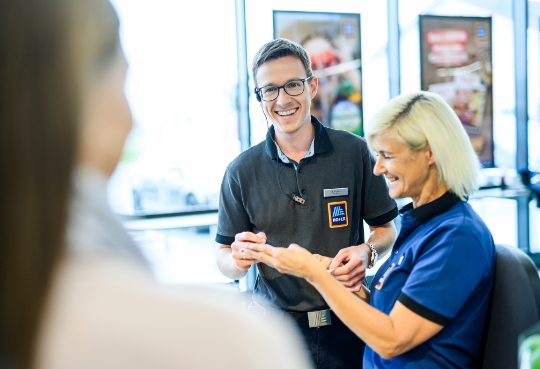 Zwei Mitarbeiter stehen in einer ALDI Suisse Filiale, beide lächeln und halten einen Kassenzettel in der Hand. Von der dritten Person sieht man nur den Hinterkopf.