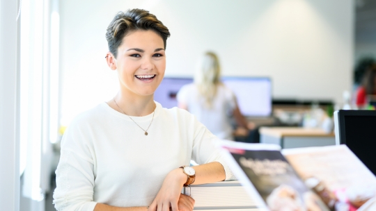 Eine Frau steht an ihrem Arbeitstisch und lächelt freundlich.