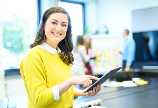 Eine Frau hält ein Tablet in der Hand und lächelt. Im Hintergrund sind Personen zu sehen die an einem Flipchart arbeiten.