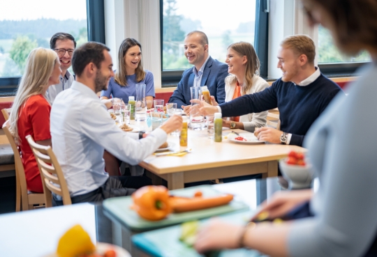8 Personen darunter Frauen und Männer sitzen an einem Esstisch und unterhalten sich freundlich und essen dabei gesunde Snacks.