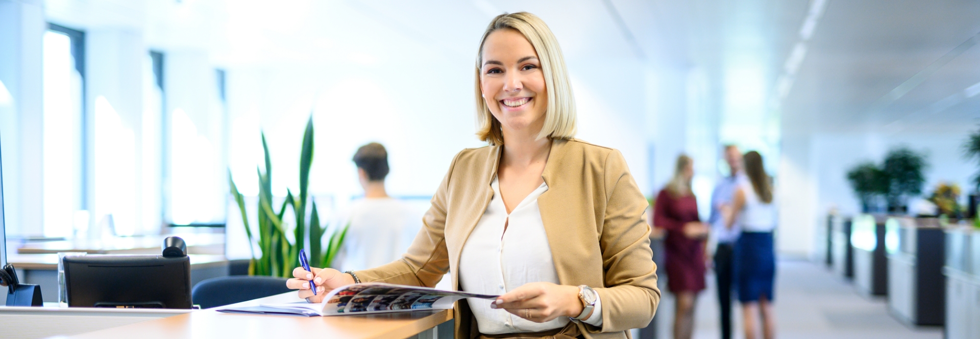 Una donna sorridente è in piedi alla reception e sfoglia un opuscolo, sullo sfondo si vedono delle persone che parlano.